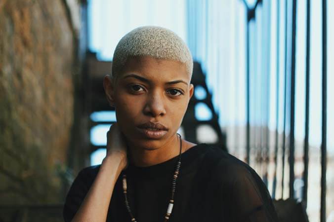 Black womxn with a professional shortly shaved and dyed blond hairstyle standing in front of a staircase looking to the camera