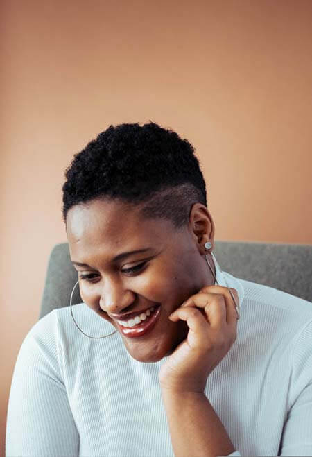 Black womxn with a professional short TWA hairstyle with shaved sides wearing hoops, sitting and smiling away from the camera