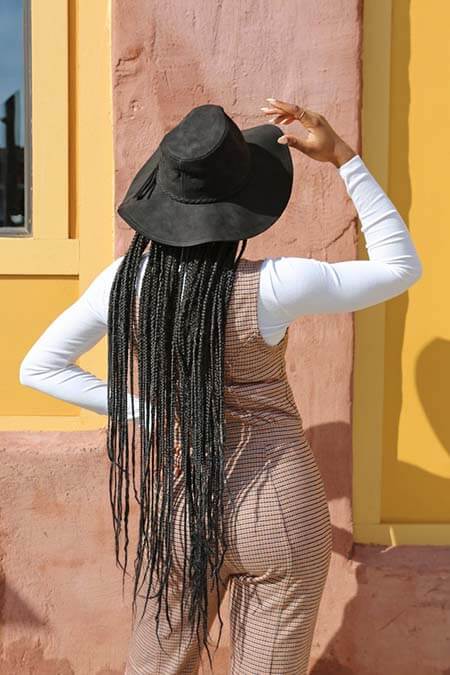 Black womxn with a professional very long ghana braids hairstyle wearing a hat and standing with her back to the camera
