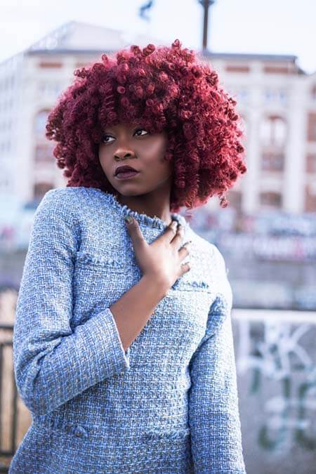 Black womxn with a professional dyed red big curls afro hairstyle wearing a blue dress standing in an urban setup