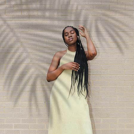 Black womxn with a professional long faux locs hairstyle wearing a pale yellow dress standing in front of a brick wall