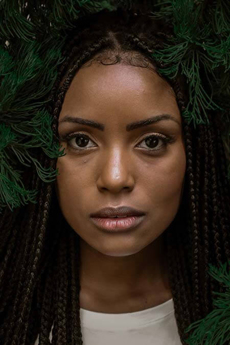A portrait of a black womxn with a professional box braids hairstyle, stands under a pine tree