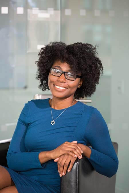 Black womxn with a professional 4C afro hairstyle wearing glasses and a blue dress, sitting on a chair