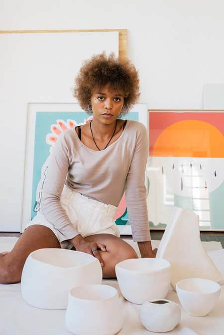 Black womxn with a professional light afro hairstyle sitting in a room next to white pottery items