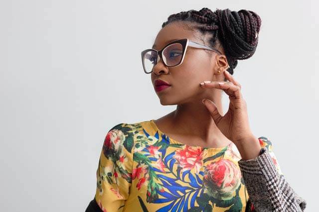 Black womxn with professional havana twists hairstyle wearing glasses in a yellow top, sitting in front of a white wall